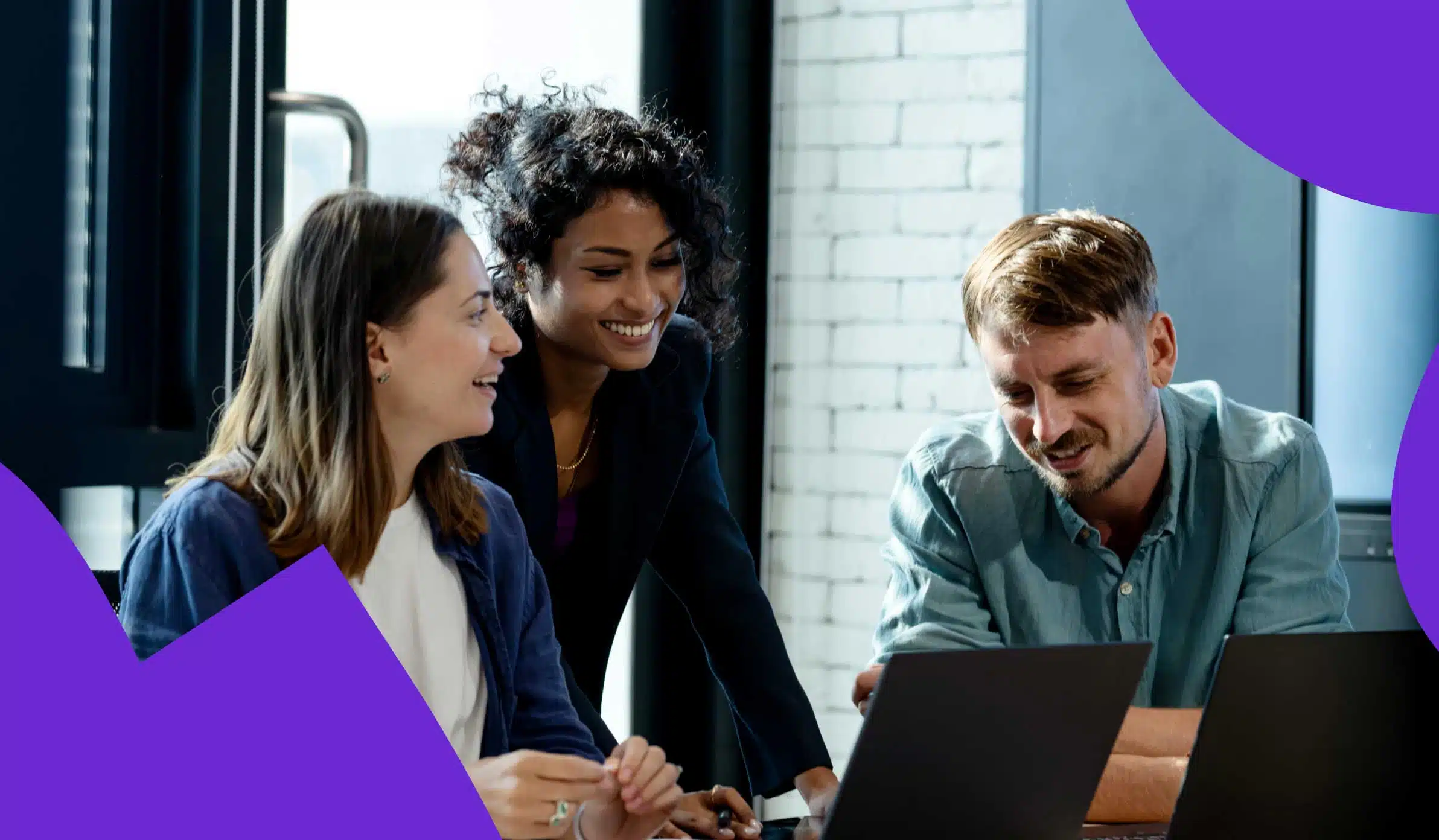 Group of three employees standing around a laptop, laughing and discussing work